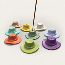 Load image into Gallery viewer, Eight pastel cowboy hat incense holders in blush, teal, yellow, mint, white, blue, olive, and purple arranged in a circle on a cream surface with a brown incense stick inserted in the center sage one
