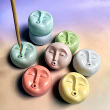 Load image into Gallery viewer, A set of eight small round ceramic incense burners with stylized sleeping face designs in pastel colors, including mint green, light blue, sage green, pale pink, white, coral pink, and pale yellow. One incense stick is shown being held into the front-left burner.
