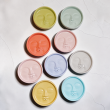 Load image into Gallery viewer, Eight round moon face incense holders in olive, mint, coral, pink, lavender, gray, yellow, and blue arranged in a scattered flat lay on a white marble surface
