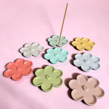 Load image into Gallery viewer, Seven pastel flower-shaped incense holders in white, mint, yellow, light blue, coral, and sage arranged in a circle on a pink surface with a wooden incense stick inserted in the light blue center one
