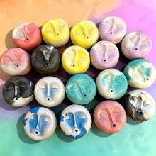 Load image into Gallery viewer, A group of small round face-shaped beads or charms arranged in rows on a pastel gradient background. The faces are painted in various colors including pink, yellow, purple, gray, blue, teal, and white, with some showing closed eyes and others with open eyes.

