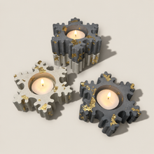 Load image into Gallery viewer, Four Christmas snowflake-shaped tealight candle holders made of textured gray concrete with scattered gold leaf accents, shown lit on a warm beige background.
