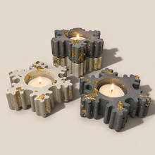 Load image into Gallery viewer, Four concrete snowflake-shaped tea light candle holders in white, light gray, and dark gray with gold leaf flake accents and lit tea lights, grouped together on a warm beige surface casting soft shadows
