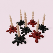 Load image into Gallery viewer, Six twisted taper candles in cream, set in black and red wax-like splatter bases, arranged on a pale pink background.
