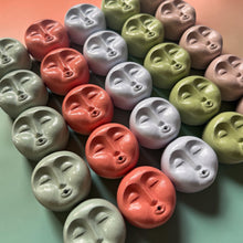 Load image into Gallery viewer, A collection of small, round, handcrafted clay figurines shaped like faces with closed eyes, shown in multiple pastel colors including sage green, coral pink, lavender, and olive green on a matching pastel background.
